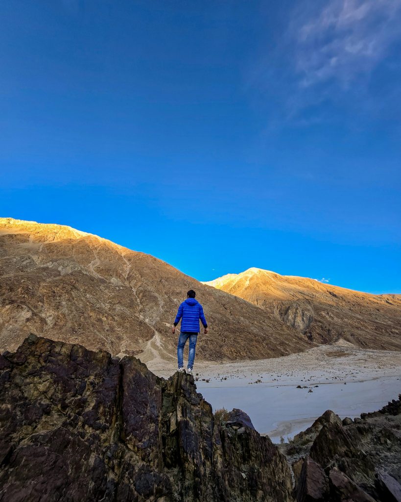 A lone traveler stands atop a rocky peak gazing at sunlit mountains under a clear blue sky.